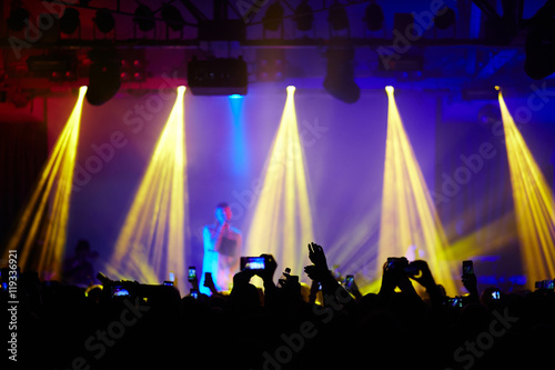 Fans in crowd raising hands up cheering in front of stage at music concert, admiring great performance of young singer in bright lights and filming it with smartphones