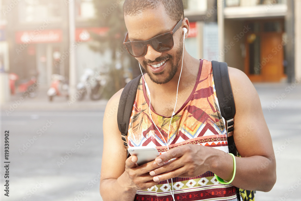 © WHstudio Leushin N - Muscular man in shades and stylish clothes, typing messages using wireless Internet connection on cell phone as walking down street, listening to new album of his favourite artist with headphones