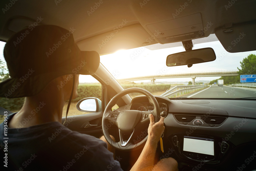 Naklejka premium Traveler in panama hat drives his car on interurban road during sunset, one hand on steering wheel and sun flares on windscreen