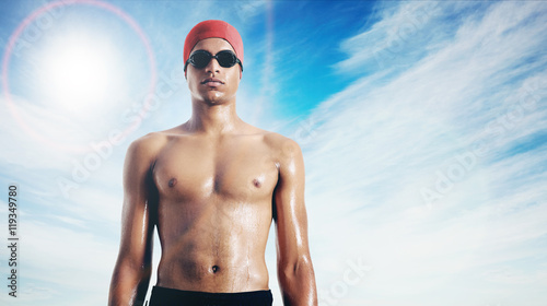 Head and torso of a serious-looking fit wet latin swimmer in red cap and black goggles against sky background with lens glare