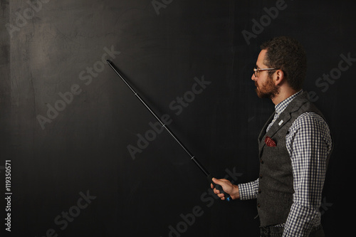 Serious bearded professor in plaid shirt and tweed vest, wearing glasses, shows something on school black board with his folding pointer