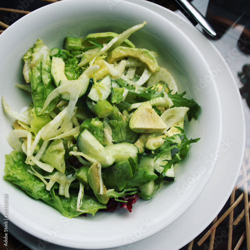 Simple green salad on the white round plate. Aged photo. Vegan dish. Raw cucumber, salad greens and avocado appetizer. Atop close up. Vegetarian Salad. Cafe photo with retro filter.