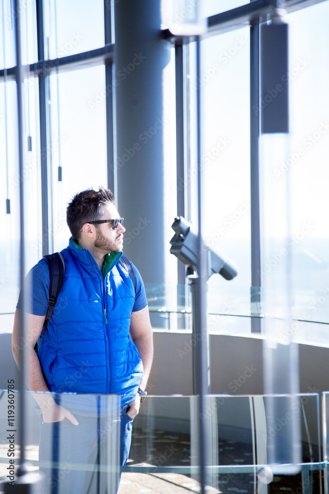 Close up of a young handsome man with black beard and modern trendy hairstyle wearing blue sleeveless jacket, jeans and sun glasses enjoying the scenic view from skyscraper. Modern interior design Close up of a young handsome man with black beard and modern trendy hairstyle wearing blue sleeveless jacket, jeans and sun glasses enjoying the scenic view from skyscraper. Modern interior design
