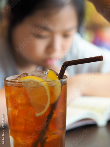 Focus on one glass of ice black tea with lemon in the coffee shop, blurred background of woman reading the book