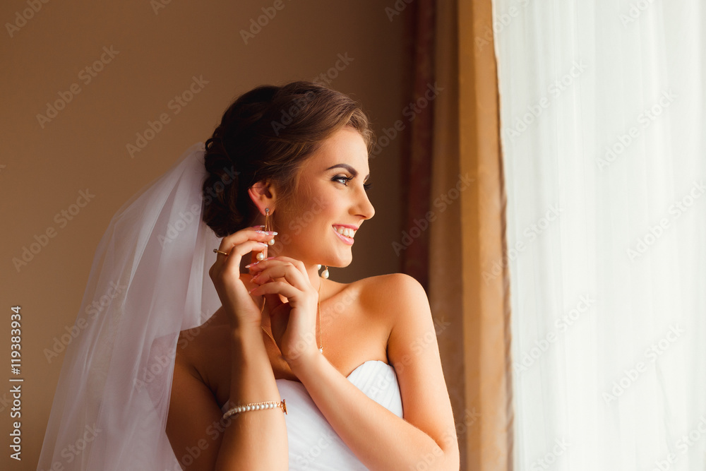 Brunette bride smiles while adjusting her earrings before the wi Brunette bride smiles while adjusting her earrings before the wi