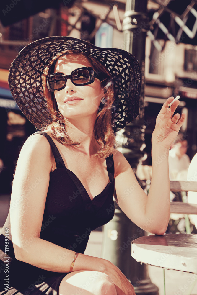 jolie femme brune style années 1960 en rob enoire et chapeau vintage fumant  sur une terrasse au soleil Stock Photo | Adobe Stock