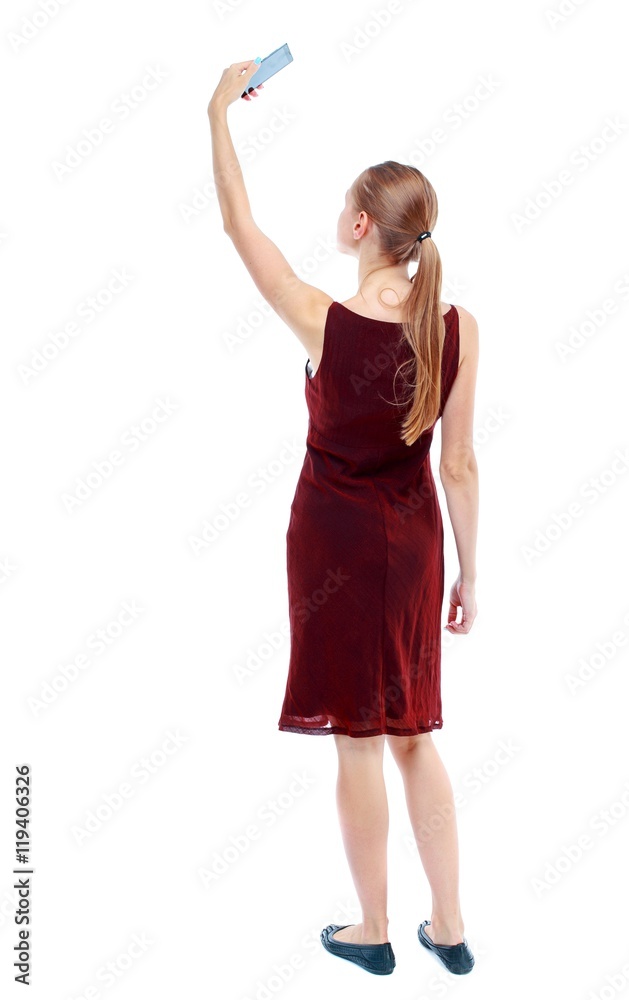 back view of standing young beautiful woman using a mobile phone. Isolated over white background. The girl in the maroon sleeveless dress makes selfie. back view of standing young beautiful woman using a mobile phone. Isolated over white background. The girl in the maroon sleeveless dress makes selfie.