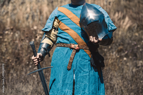 The knight in armor standing holding a sword and helmet in hand.