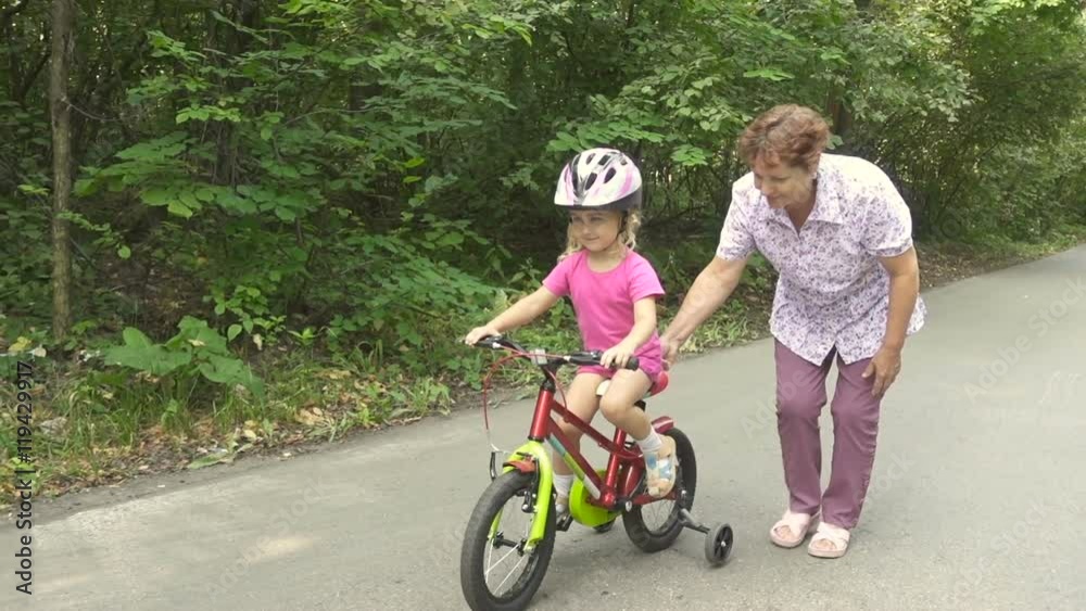 Grandmother learns to ride a bike little granddaughter. Happy child riding a bike in park outdoor. Slow motion. Grandmother learns to ride a bike little granddaughter. Happy child riding a bike in park outdoor. Slow motion.