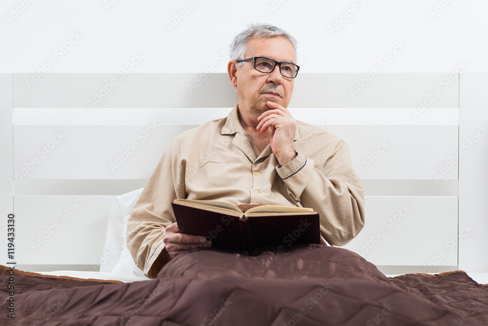Senior man is lying in bed and reading a book before sleep.