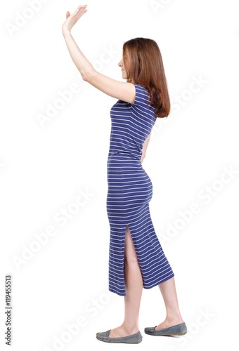Side view of beautiful woman welcomes. Young teenager girl in jeans hand waving from. Isolated over white background. The brunette in a blue striped dress stands sideways and waving his hand in © ghoststone