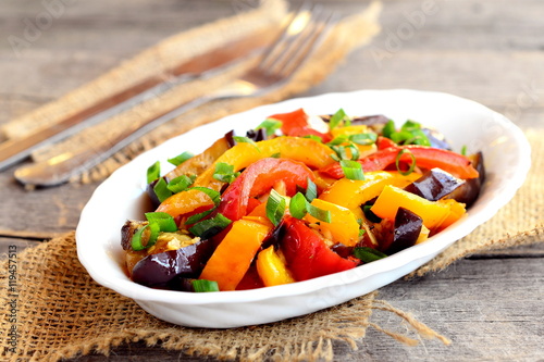 Vegetarian stew on a plate and on wooden table. Steamed aubergine, red and orange peppers and green onions. Diet dish. Vegetable stew dish. Closeup
