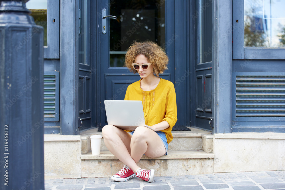 Small business. Full length portrait of a young successful woman with laptop sitting at the entrance of her shop and working online. Small business. Full length portrait of a young successful woman with laptop sitting at the entrance of her shop and working online.
