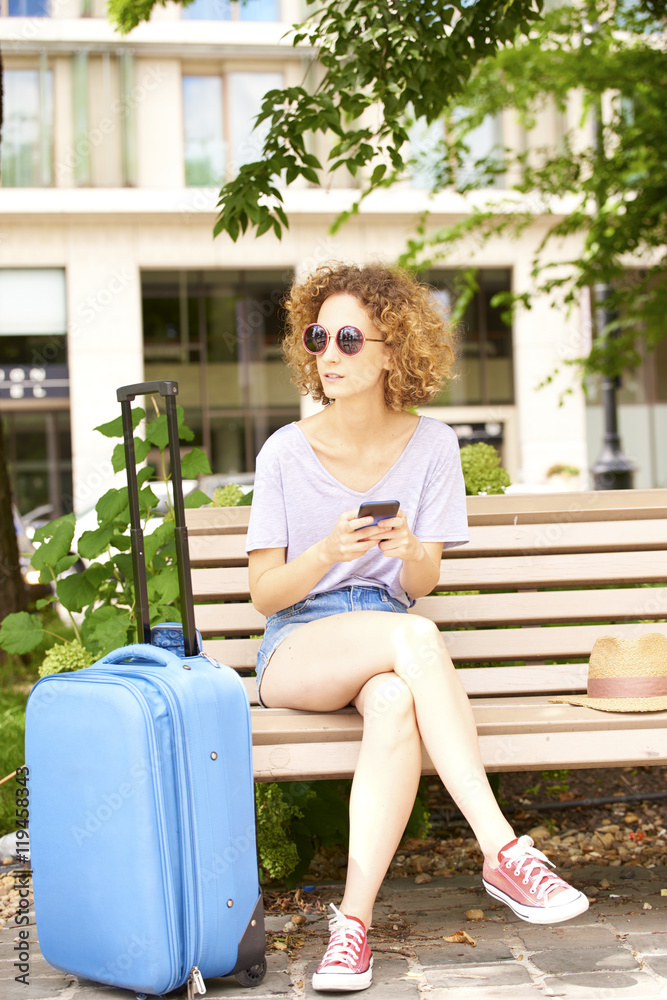 Summer in the city. Full length portrait of smiling young woman with suitcase sitting on bench and using her smartphone.