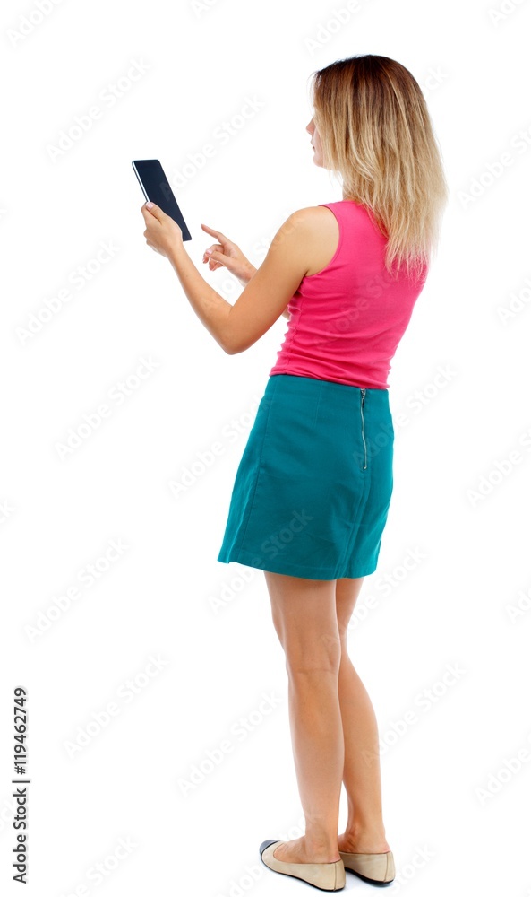 back view of standing young beautiful woman using a mobile phone or tablet computer. Isolated over white background. Blonde in a red sweater and green skirt is working on the tablet. back view of standing young beautiful woman using a mobile phone or tablet computer. Isolated over white background. Blonde in a red sweater and green skirt is working on the tablet.