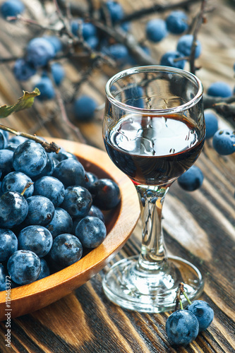 Sloe gin. Glass of blackthorn homemade light sweet reddish-brown liquid. Sloe-flavoured liqueur or wine decorated with fresh juicy ripe prunus spinosa berries on wooden background. Selective focus