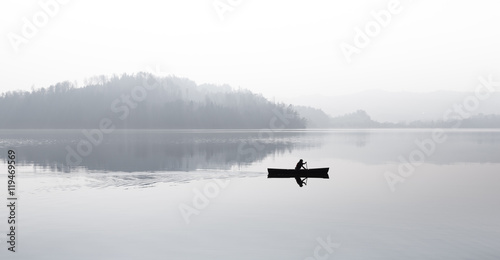 Autumn. Fog over the lake. Silhouette of mountains in the background. The man floats in a boat with a paddle.