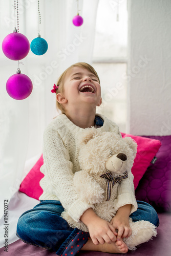 Fototapeta Naklejka Na Ścianę i Meble -  Smiling little girl sitting on pidvkonni and embraces a teddy bear. New Year's box decorated with colored balls