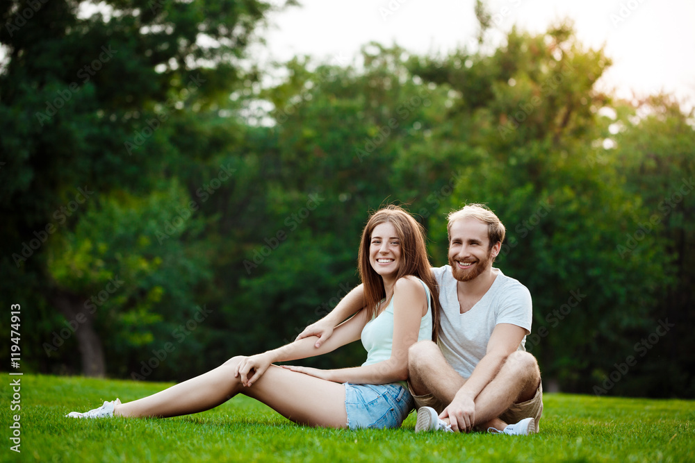 Young beautiful couple smiling, sitting on grass in park. Outdoor background. Young beautiful couple smiling, sitting on grass in park. Outdoor background.