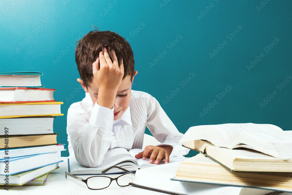 Сute boy reading difficult book at the table, blue background Сute boy reading difficult book at the table, blue background