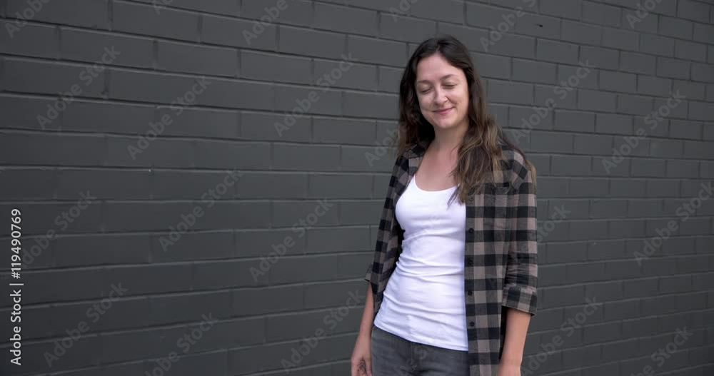 Attractive young woman in white T-shirt and plaid shirt stands next to black brick wall and smiles in Downtown LA. Medium shot, hand-held, recorded in real time at 24fps Attractive young woman in white T-shirt and plaid shirt stands next to black brick wall and smiles in Downtown LA. Medium shot, hand-held, recorded in real time at 24fps