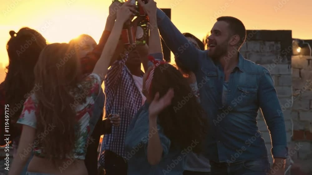 Group of multi-ethnic young people toasting with beer bottles and dancing to the music played by dj at urban rooftop party at sunset