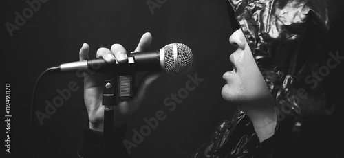 asian handsome male rocker artist in black leather hood jacket singing with dynamic microphone on dark background, bw filter