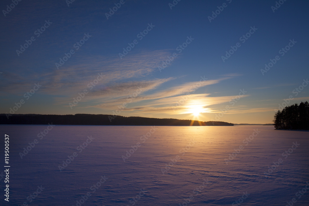 A wintry sunset. An image of a sunset on a cold winter day. Sun is going down behind a lake covered with ice and snow. Some forest is in the background. 