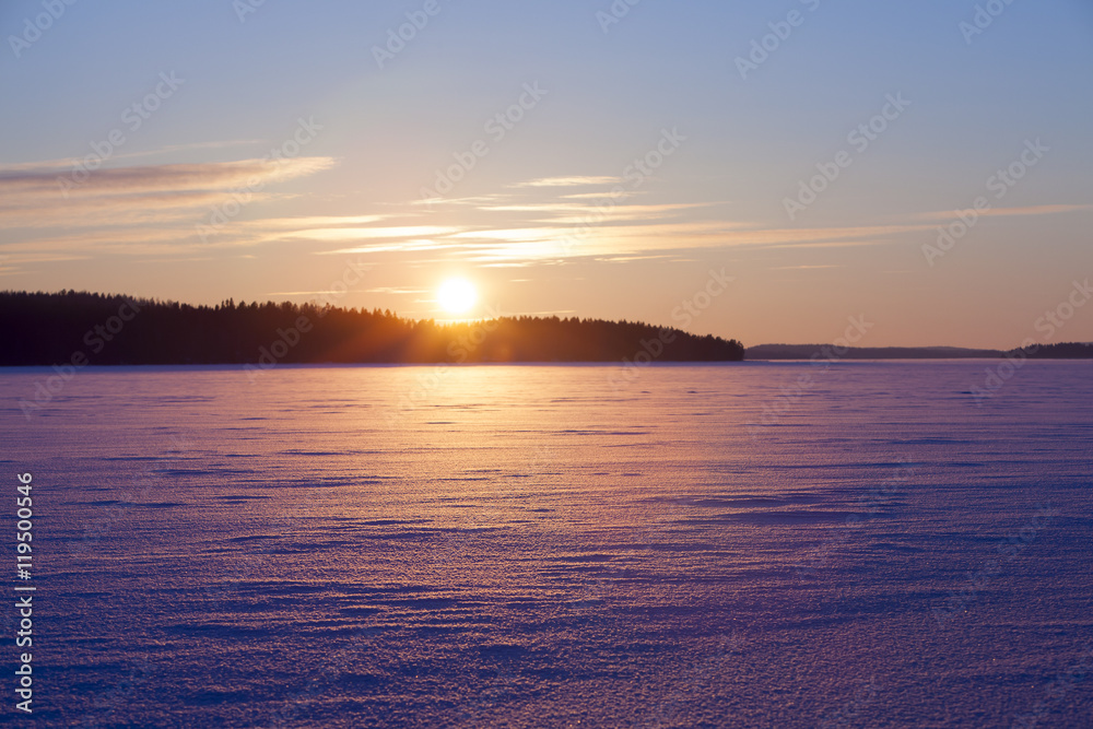 A wintry sunset. An image of a sunset on a cold winter day. Sun is going down behind a lake covered with ice and snow. Some forest is in the background. 