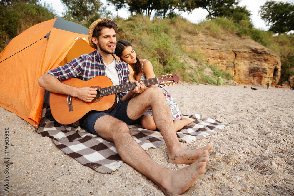Young happy couple sitting at the tent and playing guitar