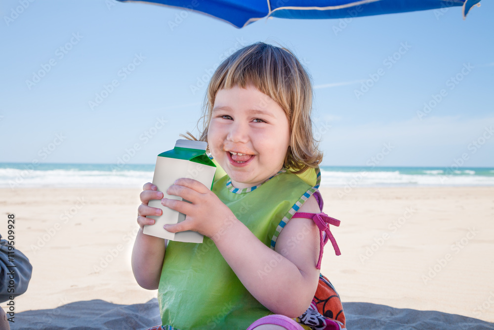 two years old child with green bib sitting down parasol umbrella at beach drinking from blank beverage container in hands looking smiling Stock Photo | Stock
