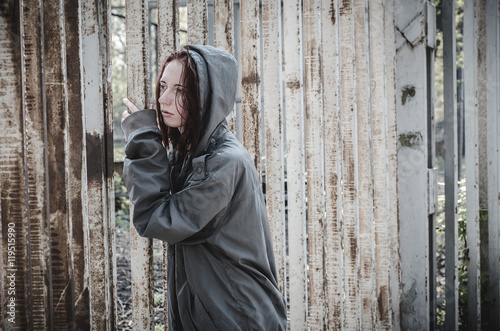 sad young girl in the old dirty clothes standing at the iron fence and sadly looks through it. Homeless devshuka. A homeless teenager.