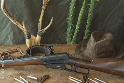 Still life. Hunting rifle, antlers, some bullets, vintage trap,belt and cowboy hat on a wooden background in front of hunter clothes