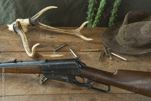 Still life. Hunting rifle, antlers, some bullets, vintage trap,belt and cowboy hat on a wooden background in front of hunter clothes