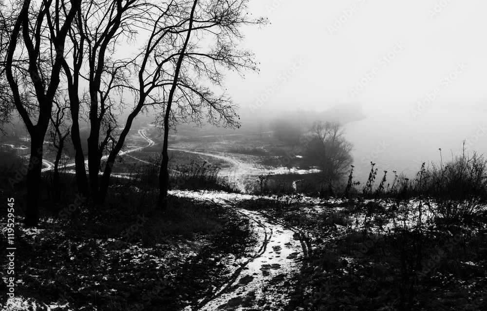 Winter landscape with frozen river. Snowy path and trees on top .of hill. Lake is covered with ice in heavy fog. Snowy weather. Black and white photo