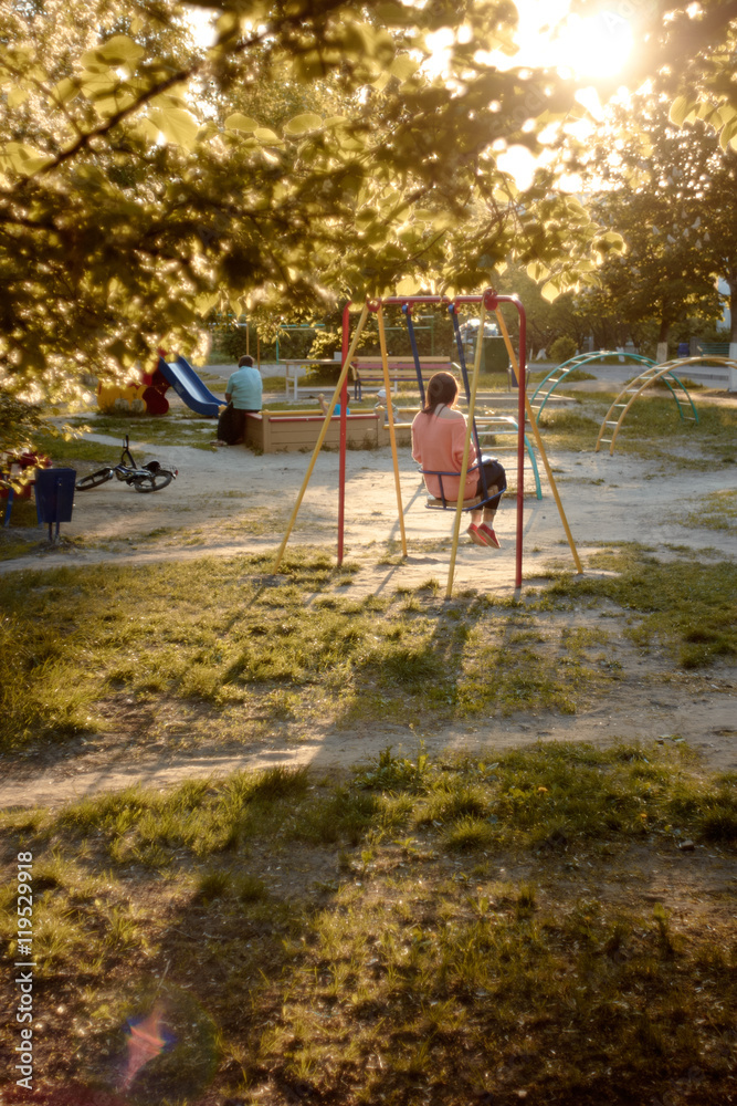 Fototapeta premium Girl on swing. Lonely girl on playground casts long shadow on grass in backlight. Foliage, illuminated by evening sun. Sad mood. loneliness