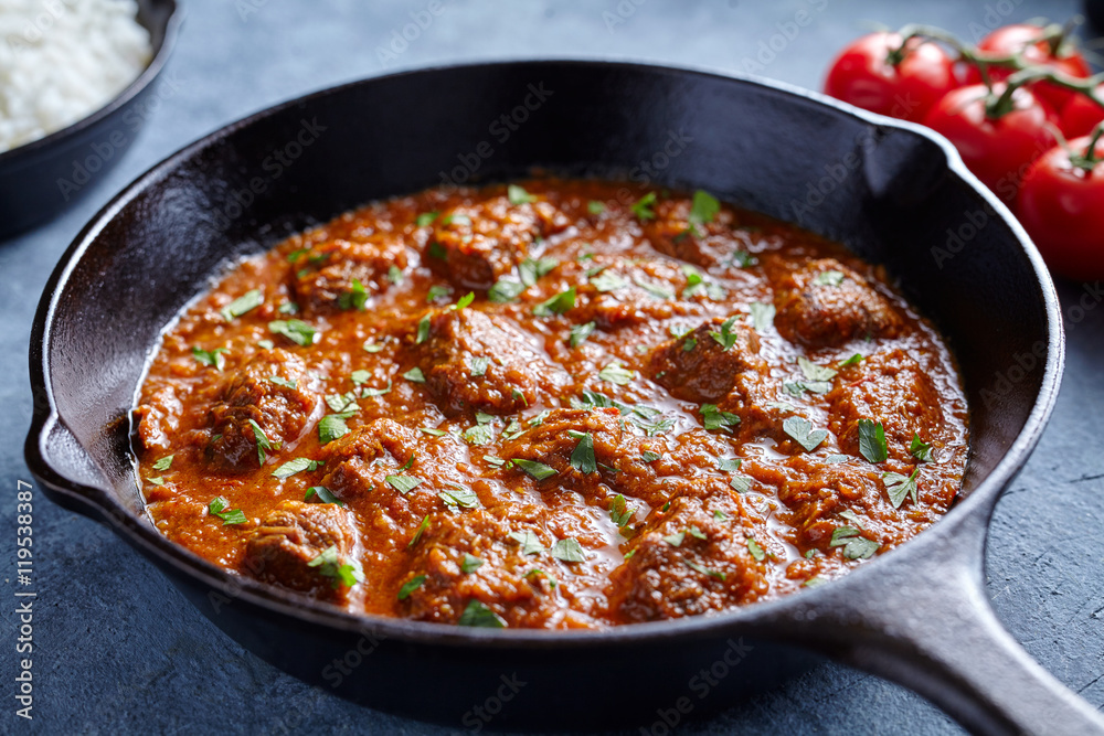 Madras butter Beef spicy slow cook lamb food with tomatoes in cast iron pan on blue table background. Delicious India culture restaurant dish.