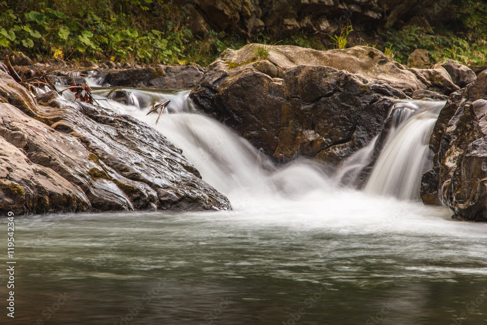 Fototapeta premium Landscape of mountain river in sunshine. View of the stony rapids. Fast jet of water at slow shutter speeds give a beautiful magic effect.