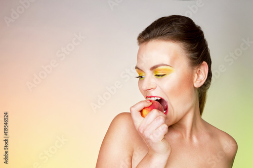 Молодая женщина с желтыми тенями на глазах эмоционально откусывает персик.