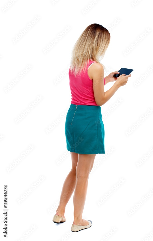 back view of standing young beautiful woman using a mobile phone or tablet computer. Isolated over white background. Blonde in a red sweater and green skirt playing tablet. back view of standing young beautiful woman using a mobile phone or tablet computer. Isolated over white background. Blonde in a red sweater and green skirt playing tablet.