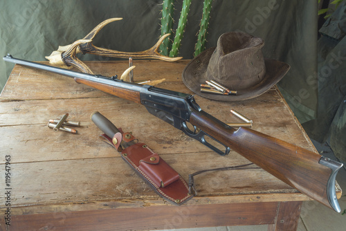 Still life. Hunting rifle, antlers, some bullets, vintage trap,belt and cowboy hat on a wooden background in front of hunter clothes