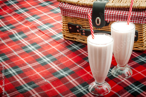 Two cold strawberry smoothies in milkshake glasses with each smoothie having a striped straw. The milkshakes are in front a wicker basket on a red picnic blanket with copy space photo