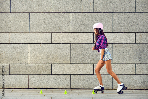 Fototapeta Naklejka Na Ścianę i Meble -  Beautiful girl rollerblading on the urban grey background in a park. Young woman on roller skates and a pink helmet. Sport lifestyle.