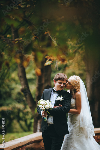 Glad newlyweds stand on fallen leaves in autumn park. Froom kissing bride. Green and red forest. photo