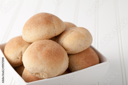 Fresh homemade whole wheat rolls on white wood in a square bowl on white wood