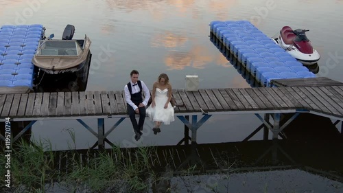 Happy new married couple sitting on dock near water reflecting sky and pink sunset clouds photo