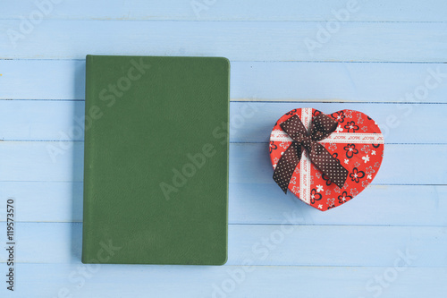 Red heart-shaped gift box and  a green book  on blue wooden background.