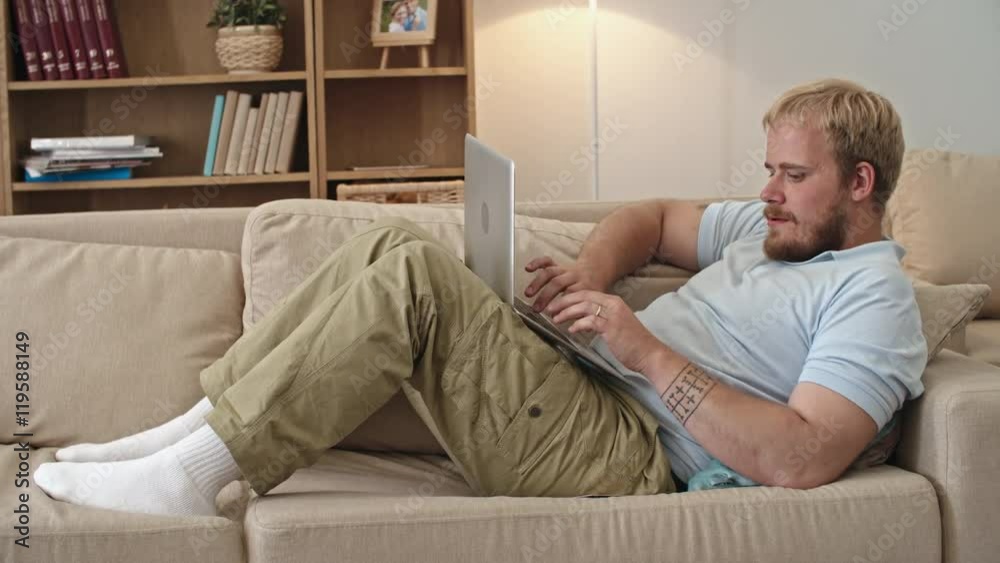 Man lying on the couch, taking credit card and typing on his laptop while shopping online Man lying on the couch, taking credit card and typing on his laptop while shopping online
