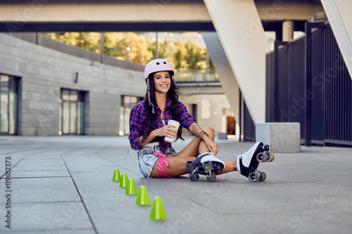 Young woman take a rest after riding roller skates and drinking coffee or tea sitting on the pavement on street. Happy girl enjoying roller skating rollerblading in urban park.