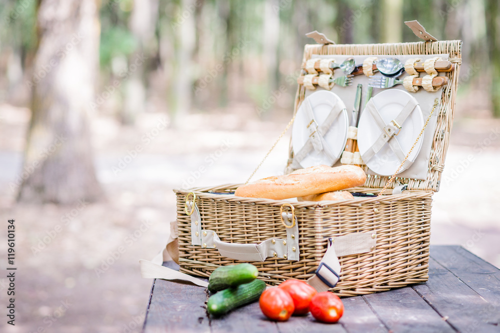 Open picnic basket with tomatoes, cucumbers and bread over a wooden table in the park.
